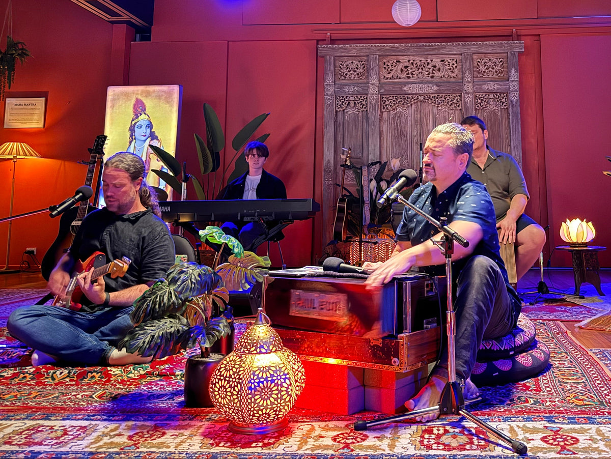 A live band performing on a decorated stage with red walls, hanging white paper lanterns, and patterned rugs. The front performer is seated at a microphone and keyboard, with other musicians playing guitar, drums, and instruments in the background.